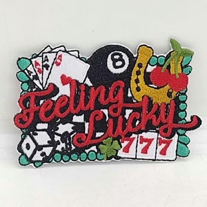 May include: An embroidered patch featuring the text Feeling Lucky in red script. The design includes white playing cards, a black eight ball, a yellow horseshoe with red cherries, white dice, a green four-leaf clover, and red 777 symbols.