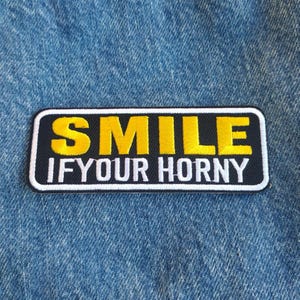 May include: A rectangular patch with the words "SMILE IF YOUR HORNY" in yellow and white lettering on a black background. The patch is bordered with a white outline and is set against a blue denim fabric.
