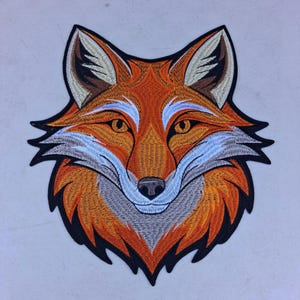 May include: Embroidered patch featuring a detailed fox head. The fox has orange, white, gray, and black colors. The patch is designed with intricate stitching, showcasing the fox's features, including its eyes, nose, and fur.