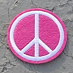 May include: A pink peace sign patch with a white outline. The patch is made of felt and has a fuzzy texture.