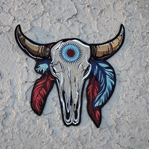 May include: A white embroidered bull skull with blue, red, and gold feathers. The skull has a blue and white circle with a red center in the middle of the forehead.