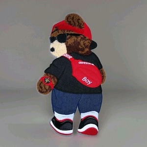 May include: A brown teddy bear patch wearing a red baseball cap, black sunglasses, a black t-shirt, blue pants, and black and red sneakers. The bear has a red crossbody bag with the white text "awesome Boy" and a red watch on its left arm.