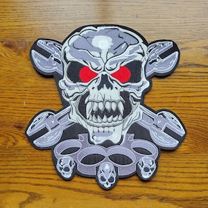 May include: A gray skull with red eyes, crossed wrenches, and a silver knuckle duster. The skull has a menacing expression with sharp teeth.