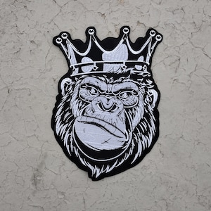 May include: Black and white embroidered patch featuring a gorilla wearing a crown. The gorilla has a stern expression, with detailed facial features. The crown is also black and white, with a simple design. The patch is likely for clothing or accessories.