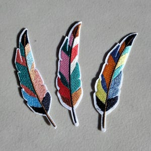 May include: Three colorful embroidered feather patches. Each patch features a different color scheme, including teal, orange, pink, and blue, with a white border and black accents. These patches are ideal for adding a decorative touch to clothing or accessories.