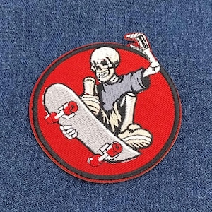 May include: A red and black circular embroidered patch featuring a skeleton skateboarding. The skeleton is wearing a grey shirt and is holding a skateboard with red wheels.