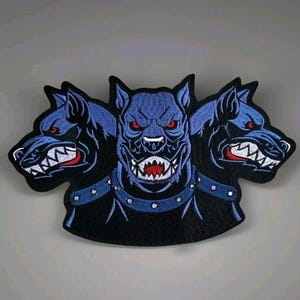 May include: Embroidered patch featuring a three-headed dog with blue fur, red eyes, and bared teeth. The patch has a black border and a blue collar with silver studs. The dog's snarling expression and menacing appearance create a bold design.