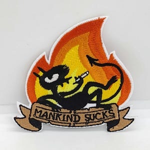 May include: Embroidered patch featuring a black cartoon devil reclining in front of a flame. The devil is holding a cigarette. A banner below reads "MANKIND SUCKS" in brown letters.