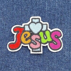 May include: A colorful embroidered patch with the word "Jesus" in rainbow colors and a white heart on a cross.