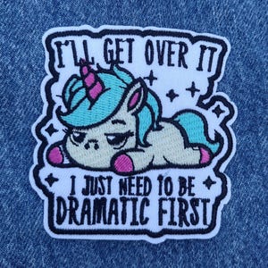 May include: Embroidered patch featuring a cartoon unicorn with a turquoise mane and pink accents, lying down with a bored expression. The patch includes the text "I'LL GET OVER IT, I JUST NEED TO BE DRAMATIC FIRST" in black lettering.