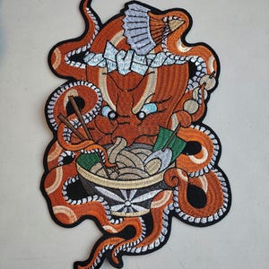 May include: Embroidered patch featuring an orange octopus with a stern expression, wearing a crown and eating ramen from a bowl. The patch has a black border and includes details like chopsticks and green garnish.