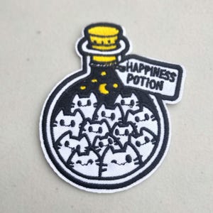 May include: Embroidered patch featuring a potion bottle filled with white cat faces. The bottle has a yellow stopper and the text "HAPPINESS POTION" on a white label. The background inside the bottle is black with a yellow crescent moon and stars.