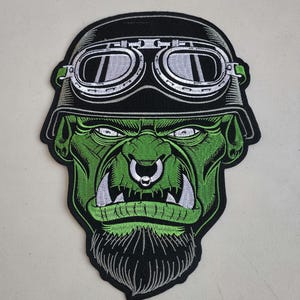 May include: Green and black embroidered patch of a green orc wearing a black helmet with goggles and a nose ring.