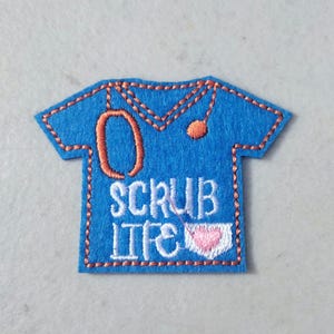 May include: A blue felt patch in the shape of a medical scrub top. The patch features an orange stethoscope design and the words "SCRUB LIFE" in white. A pink heart is also included in the design. The patch is outlined with orange stitching.