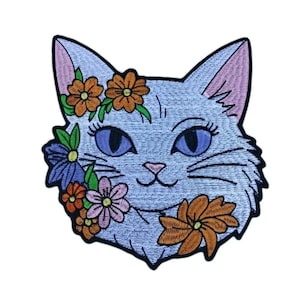 May include: Embroidered patch featuring a light blue cat face adorned with orange, pink, and purple flowers. The cat has blue eyes, pink ears, and a smiling expression. The patch is designed with a detailed, stitched appearance.