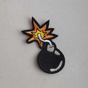 May include: Embroidered patch featuring a cartoon bomb with a black body and a white fuse. The explosion is depicted with yellow and orange flames, outlined in black. The patch is designed for application on clothing or accessories.