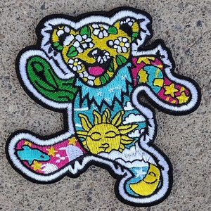 May include: A colorful embroidered patch featuring a cartoon bear with a sun, moon, and flowers. The bear is wearing a blue, green, and pink patterned shirt.