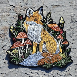 May include: Embroidered patch featuring a fox sitting in a forest setting. The fox is orange and white with a bushy tail. The background is green with mushrooms and flowers.