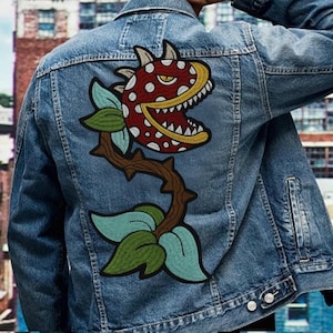 May include: A blue denim jacket with a large embroidered patch of a red, white, and yellow plant with a toothy mouth. The plant has green leaves and a brown stem with thorns.