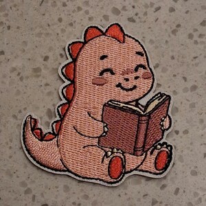 May include: An embroidered patch featuring a light orange dinosaur with red spikes along its back and tail. The dinosaur has a smiling expression and is holding an open brown book with both hands, showcasing detailed stitching and a white border.