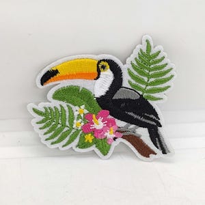 May include: Embroidered patch featuring a toucan with a yellow and orange beak, black body, and white accents. The toucan is perched on a brown branch with pink flowers and green leaves. The patch has a white border.