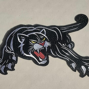 May include: Black panther patch with white and gray accents. The panther is snarling, with its mouth open, revealing a red tongue and white teeth. The eyes are yellow. The patch is embroidered with a detailed design.