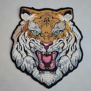 May include: Embroidered tiger head patch with a black border. The tiger features orange and white fur, green eyes, and a pink mouth with visible teeth. The patch is designed to be sewn or ironed onto fabric.