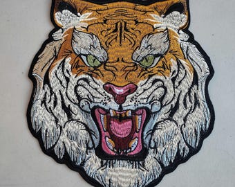 May include: Embroidered tiger head patch with a black border. The tiger features orange and white fur, green eyes, and a pink mouth with visible teeth. The patch is designed to be sewn or ironed onto fabric.