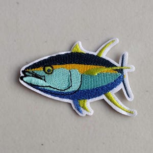 May include: Embroidered patch of a tuna fish. The fish has a blue, yellow, and black color scheme with a white border. The patch is designed to be ironed on or sewn onto fabric, suitable for clothing or accessories.