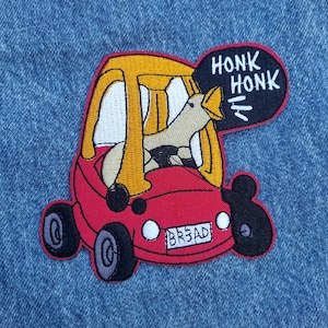May include: Embroidered patch featuring a cartoon duck driving a red car. The car has yellow trim and black wheels. The duck is honking, with the words "HONK HONK" in a speech bubble. The license plate reads "BR3AD".