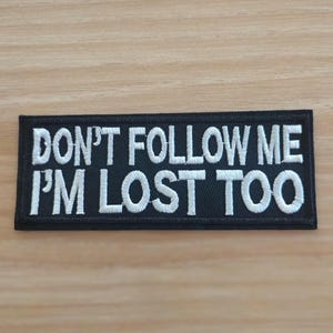 May include: A rectangular black embroidered patch with a white border. The patch features the white embroidered text DON'T FOLLOW ME I'M LOST TOO in a bold, sans-serif font, arranged in two lines across the center of the black fabric background.