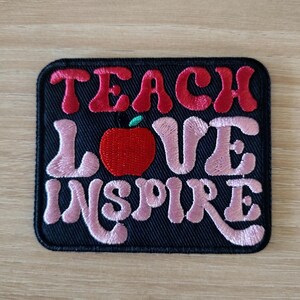 May include: Embroidered patch with the words "TEACH LOVE INSPIRE" in red and pink lettering. A red apple with a green leaf is in the center. The patch is rectangular with rounded corners and a black background.