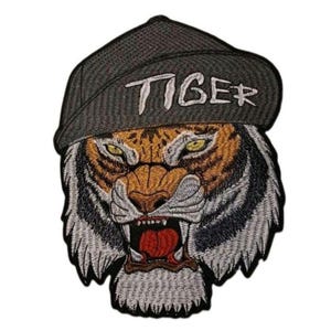 May include: An embroidered patch featuring a tiger head with yellow eyes and an open mouth showing red tongue and teeth. The tiger wears a dark gray baseball cap with the word TIGER stitched in white thread across the front of the cap brim.