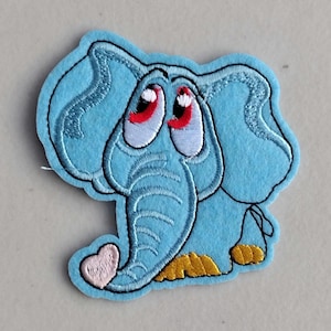 May include: A light blue felt elephant patch with embroidered details. The elephant has large, expressive eyes with red pupils, a trunk, and a heart-shaped tail. The patch has yellow feet and a black outline.