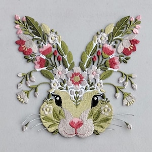 May include: Embroidered bunny face with floral accents. The bunny's face is a pale yellow-green, with black eyes and a pink nose. The ears and head are adorned with green leaves, pink and white flowers, and delicate white whiskers. A charming, spring-themed design.