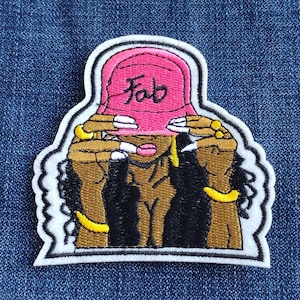 May include: Embroidered patch featuring a stylized figure wearing a pink hat with the word "Fab" in black script. The figure has brown skin, black hair, and gold jewelry. The patch is outlined in white and set against a denim background.