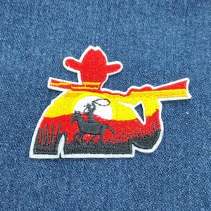 May include: Embroidered iron-on patch featuring a silhouette of a cowboy riding a horse with a lasso in his hand, set against a sunset backdrop with red, yellow, and orange colors. The cowboy is wearing a red hat.