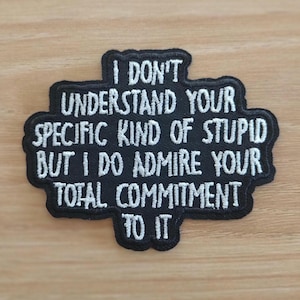 May include: Black embroidered patch with white text that reads: "I DON'T UNDERSTAND YOUR SPECIFIC KIND OF STUPID BUT I DO ADMIRE YOUR TOTAL COMMITMENT TO IT." The patch has a black border and is on a light brown surface.