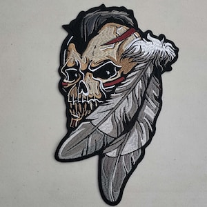 May include: Embroidered patch featuring a skull with a fierce expression, adorned with feathers. The skull is depicted in shades of brown, beige, and red, while the feathers are a light gray. The patch is set against a black background.