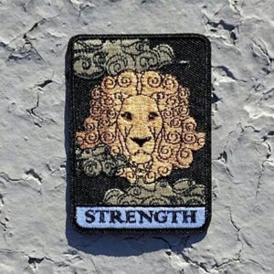 May include: A black embroidered patch with a lion's head in the center. The lion is surrounded by clouds and the word "STRENGTH" is printed at the bottom.