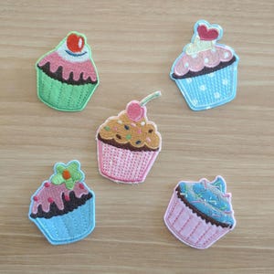 May include: Five embroidered cupcake patches in various colors. Each patch features a different design, including cherries, sprinkles, and floral accents. The cupcakes have green, pink, and blue frosting and liners. These patches are ideal for decorating clothing or accessories.