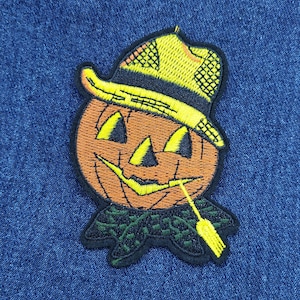 May include: Embroidered patch featuring a smiling jack-o'-lantern wearing a yellow straw hat. The pumpkin has black eyes and a stitched mouth with a straw. The patch has a black border and green collar, perfect for Halloween.