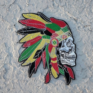 May include: Embroidered patch featuring a skull wearing a Native American headdress. The headdress has feathers in red, yellow, green, and black. The skull is white with detailed shading. The patch is likely for clothing or accessories.