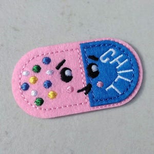 May include: A pink and blue felt pill-shaped patch with embroidered details. The pink side has colorful embroidered dots and a smiling face. The blue side says "CHILL" in white embroidery, also with a smiling face. The patch has a stitched border.