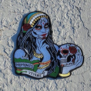 May include: Embroidered patch featuring a blue-skinned woman with a skull. The woman wears a headscarf, jewelry, and a rose. The skull has a red rose and decorative details. The patch includes the text "MEMENTO VIVERE".