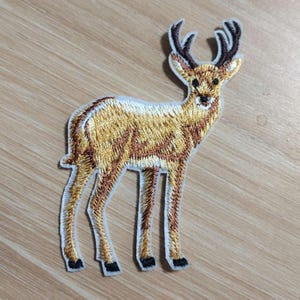 May include: Embroidered patch featuring a deer with brown and gold fur, black hooves, and dark brown antlers. The patch has a white border and is designed to be ironed or sewn onto fabric. The deer is standing.
