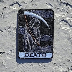 May include: Black and white embroidered patch with a skeletal figure holding a scythe. The patch has a white border and the word "DEATH" in white lettering.