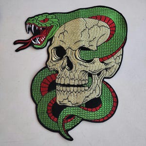 May include: Embroidered patch featuring a skull entwined by a green snake with red accents. The snake's head is open-mouthed, revealing sharp white teeth and a red tongue. The skull is off-white with detailed shading.