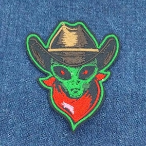 May include: Green alien wearing a brown cowboy hat with a red bandana. The alien has large eyes and a green face. The patch is embroidered and has a green outline.