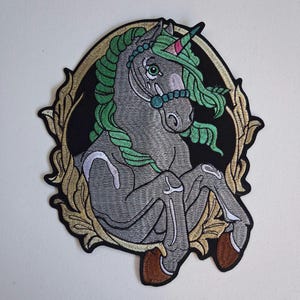 May include: An embroidered patch featuring a grey unicorn with a green mane and tail, a teal bridle, and a pink-tipped horn. The unicorn is centered within an oval frame with gold-colored ornate leaf detailing against a solid black background.